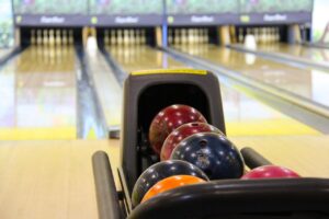 A photo of a bowling ball return with multiple different colored bowling balls in it, looking out over four bowling lanes, which are blurred in the background.