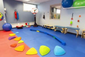 Photo of Helen Hayes Hospital pediatric rehabilitation suite. The room has red and blue matting on floor and there are a variety of colorful pieces of therapy equipment, including blue and green balls, ablue balance beam, red, purple, yellow, orange, green, and blue foam triangles, a basketball hoop, and more. 
