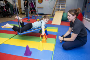 Young pediatric patient lies on her stomach on a flat swing platform and places an orange hoop around an orange cone, while therapist looks on, smiling. 