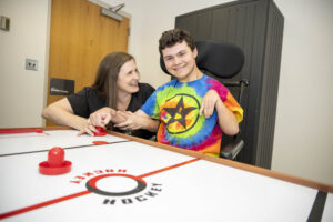Physical therapist and pediatric patient in motorized wheelchair sit smiling at an air hockey table, both with their hands on the same air hockey paddle, as therapist supports patient playing air hockey. 