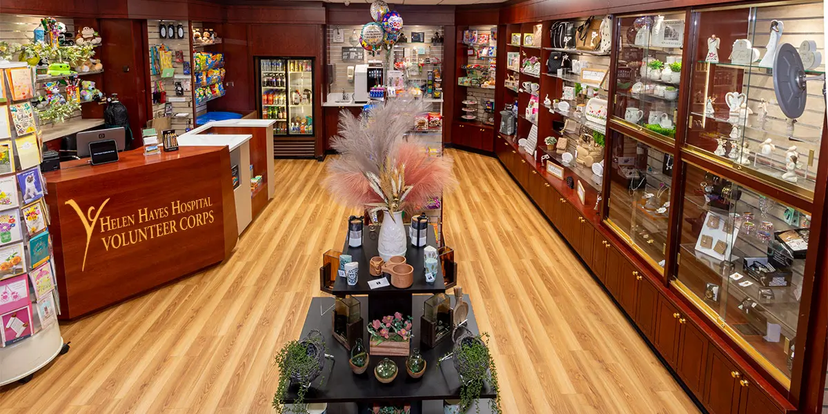 A photo of the Helen Hayes Hospital Gift Shop. A counter on the left has the writing "Helen Hayes Hospital Volunteer Corps." There is a card display to the left of the counter, snacks, a refrigerator, and a coffee machine at the back of the gift shop, and gifts such as mugs, jewelry, and household decor on the right side of the gift shop, all housed in cheery wood shelves. At the center front of the gift shop is another display with household decor for sale, such as artificial succulents and flowers.
