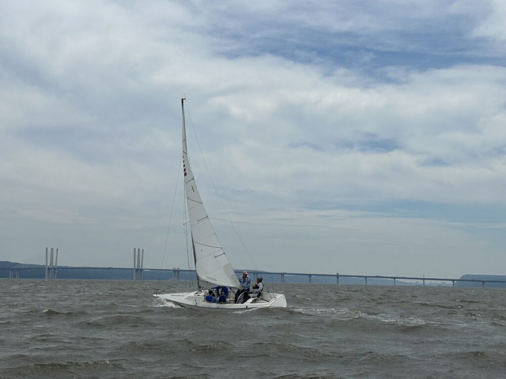 A sailboat from the Nyack Boat Club's Sonar Fleet 23 sailing on the Hudson River with tree covered mountains in the background and a blue and cloudy sky above. The boast is heading right with participants seated inside the boat.