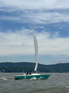 A sailboat from the Nyack Boat Club's Sonar Fleet 23 sailing on the Hudson River with tree covered mountains in the background and a blue and cloudy sky above. The boast is heading right with participants seated inside the boat.