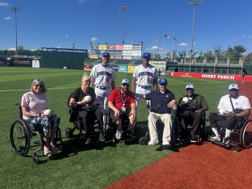 Participants in Helen Hayes Hospital's Adapted Sports & Recreation Program's Adaptive Baseball Clinic with the New York Boulders pose in their wheelchairs on the field of Boulder's Stadium with two Boulders baseball players standing behind them.