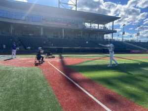 A woman sits on her rolling walker at home plate batting with a baseball bat as a player from the New York Boulders pitches a 16 inch adapted softball to her and a Boulders player stands behind her as catcher.