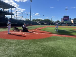 A man in a power wheelchair sits at home plate batting with a baseball bat as a player from the New York Boulders pitches a 16 inch adapted softball to him and a Boulders player stands behind him as catcher.